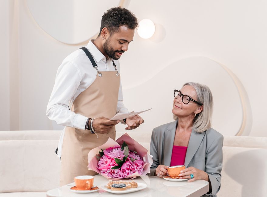 A young male florist with dark, curly hair, wearing a white shirt and beige apron, hands a floral gift note to an older woman with gray hair and glasses, who is seated at a table. The table holds a bo