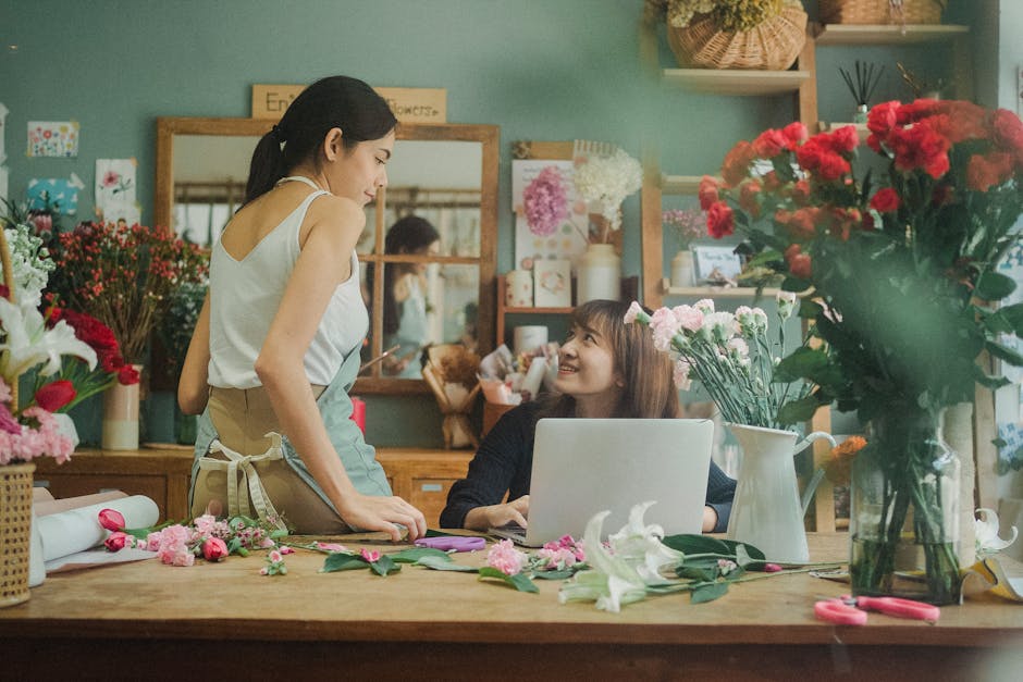 A florist and a customer share a moment in a flower shop, surrounded by vibrant flower arrangements. The florist, wearing a light-colored apron, leans over a wooden worktable, engaged in floral design