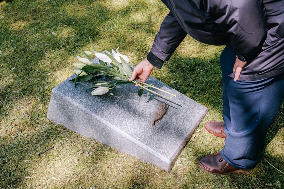 A person dressed in dark clothing and brown shoes placing a floral tribute of white lilies and greenery on a rectangular granite headstone at a grave site during daytime. The flowers are arranged with