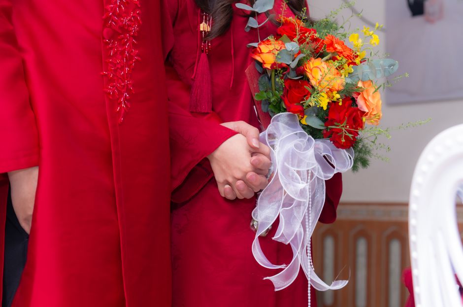 A person dressed in a red outfit holding a vibrant floral bouquet featuring red, orange, and yellow roses, complemented by greenery and small yellow filler flowers. The bouquet is wrapped with a large