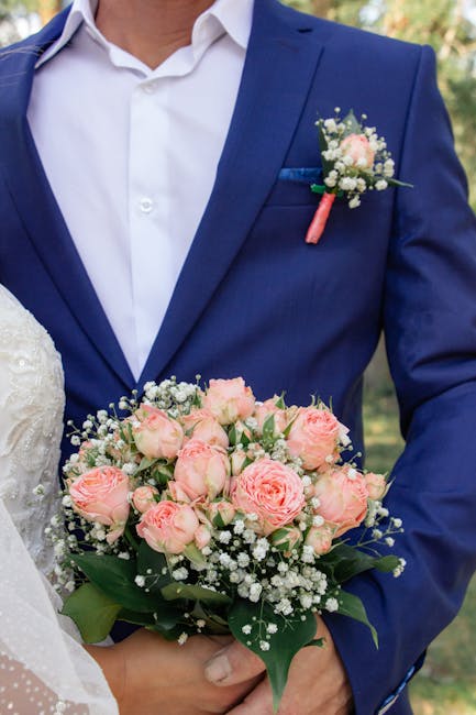 Close-up of a groom dressed in a navy blue suit with a white shirt, holding a bouquet of fresh pale pink roses, white baby's breath, and green foliage. The bouquet is arranged in a rounded style and w