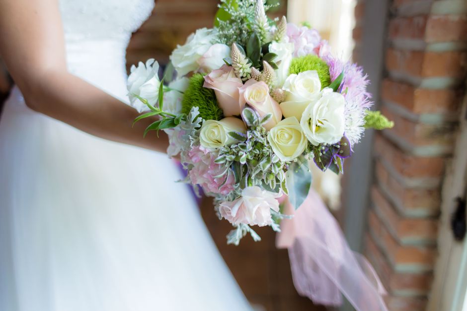 A bride holding a floral bouquet arrangement featuring creamy white roses, soft pink hydrangeas, lavender lisianthus, and green foliage, wrapped with pale pink ribbon. The bouquet is held in front of 
