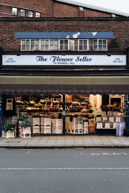 Exterior view of The Flower Seller of Muswell Hill, a flower shop with a black awning and a sign featuring blue cursive lettering, situated on a brick building facade. In the shopfront, an array of fr