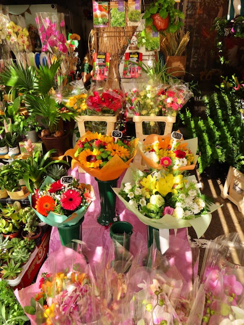 A vibrant display of fresh floral bouquets arranged on a pink table in a florist shop. The bouquets feature bright blooms such as pink gerbera daisies, yellow sunflowers, white chrysanthemums, and mix