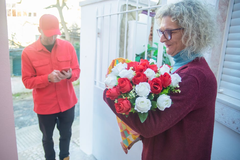 A cheerful woman with curly gray hair, wearing glasses and a burgundy sweater over a blue turtleneck, holds a vibrant bouquet of fresh red and white roses wrapped in patterned paper. She stands outdoo