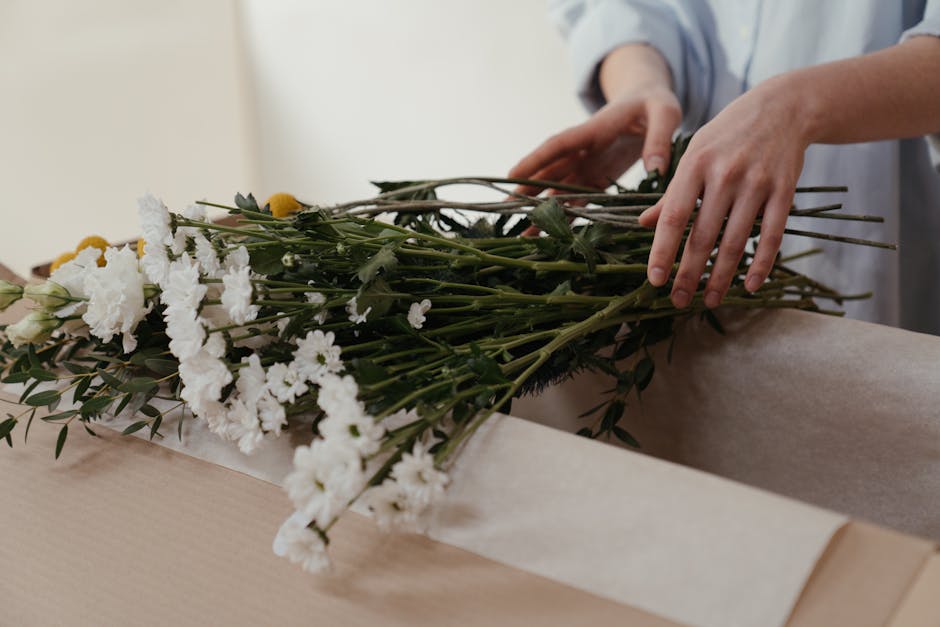 A person dressed in a light-colored shirt is placing a fresh floral bouquet wrapped in plain brown paper onto a table. The bouquet features white carnations, yellow daisies, and greenery with slender
